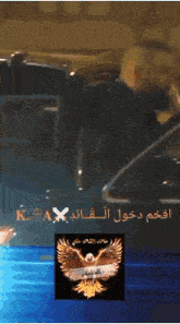 a man with a beard and a picture of an eagle with arabic writing behind him