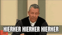 a man in a judge 's robe says hierher hierher hierher in front of a microphone