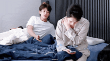 two young men are sitting on a bed and one of them is looking at his phone