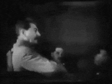 a black and white photo of a man with a mustache talking to another man in a dark room .
