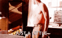 a shirtless man is standing next to a bed with the words `` just a little '' written on it .