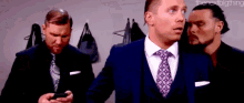 three men in suits and ties are standing next to each other in a locker room . one man is looking at his phone .