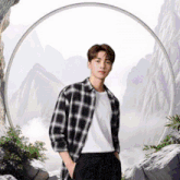 a young man wearing a plaid shirt and a white shirt stands in front of a mountain