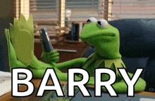 kermit the frog is sitting at a desk holding a cell phone and the word barry is on the table in front of him
