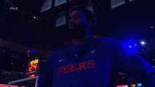 a man wearing a 76ers jacket stands in front of a scoreboard