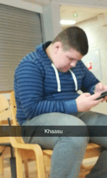 a man in a blue striped shirt sits in a chair looking at his phone and the word khaasu is on the bottom