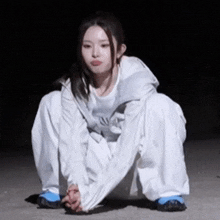 a young woman in a white jacket and white pants is squatting down in the dark .
