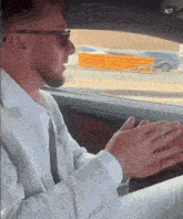a man in a suit and sunglasses is sitting in a car with his hands folded in front of him
