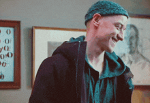 a man wearing a beanie and a jacket smiles in front of a framed picture of a man 's face