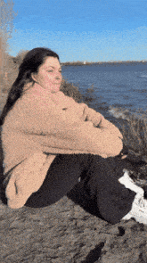 a woman in a teddy bear jacket sits on a rock near the water