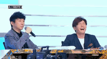 two men are sitting at a table and one is holding a microphone and the other is laughing .