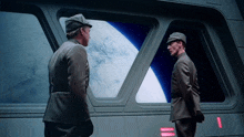 two men standing next to each other in front of a window with the earth in the background