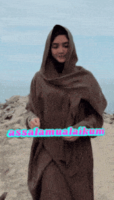 a woman wearing a hijab stands on a beach holding a sign that says ' asalamualaikum '