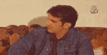 a man in a denim jacket is sitting on a couch looking at something .