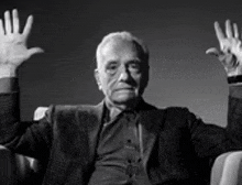 a man in a suit and tie is sitting in a chair with his hands up .