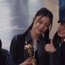 a woman in a black jacket is holding a gold statue of a man and giving a thumbs up