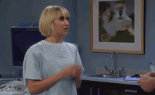 a woman in a hospital gown is standing in a room with a sink and a painting on the wall .