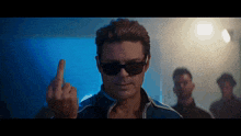a man wearing sunglasses and a blue jacket gives the middle finger