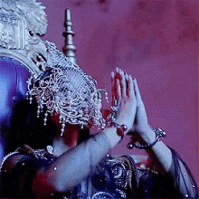 a woman wearing a mask and jewelry is sitting on a throne with her hands together .