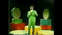 a man in a green suit is singing into a microphone on a stage surrounded by geometric shapes .