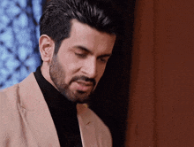 a man with a beard is wearing a tan suit and black turtleneck .