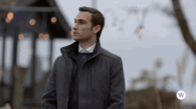 a man wearing a grey jacket and a blue sweater stands in front of a sign that says ' w ' on it