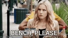 a woman is sitting at a table with a drink in her hand and the words bench please written on the screen behind her .