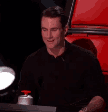 a man in a black shirt is sitting in front of a red chair and pressing a button .