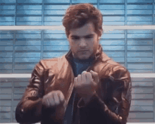 a man in a leather jacket is standing in front of a wall and holding his hands together .
