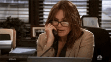 a woman wearing glasses is talking on a phone while sitting at a desk in front of a nbc logo