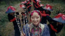 a woman with pink hair and red eyes is smiling in front of a group of people wearing red hats