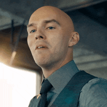 a bald man wearing a vest and tie looks to the side