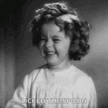 a little girl is smiling and licking the spoon .