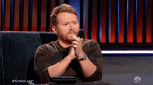 a man with a beard is sitting on a couch with his hands folded in front of a nbc logo