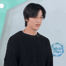 a young man in a black sweater stands in front of a sign that says debut 's plan