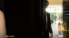 a man in a suit is standing in a dark room with a nbc logo on the bottom right