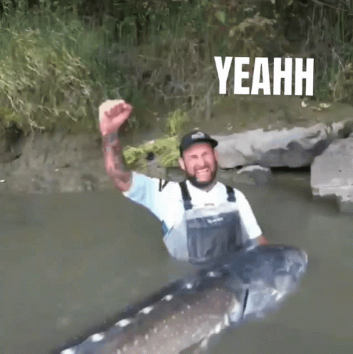 a bearded man stands in the water with a great big sturgeon fist pumped in the air "yeah!"