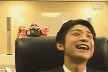 a man in a suit is laughing in front of a robot that says ' arashi spot ' on the wall behind him