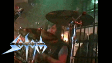 a man playing drums in front of a sign that says symo
