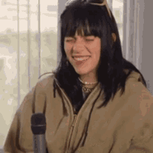 a woman in a tan jacket is smiling while holding a microphone in front of a window .