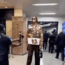 a man in a floral shirt is standing in a hallway holding a water bottle while being interviewed .