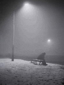 a person is sitting on a bench in the snow under a street light at night .