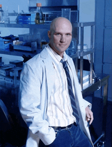 a bald man in a lab coat and tie stands in front of a shelf with gloves and a bottle on it