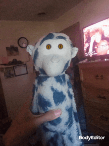 a blue and white stuffed animal with yellow eyes is being held by a person in front of a tv screen that says bodyeditor