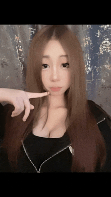 a woman with long brown hair and a black top is pointing at her face