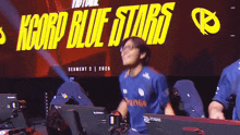 a man wearing a blue shirt that says orange is dancing in front of a sign that says kcorp blue stars