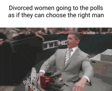 a man in a suit and tie is sitting in front of a wrestling ring holding a sign .
