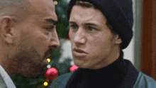 two men are looking at each other in front of a christmas tree . one of the men is wearing a black hat .