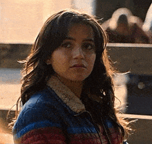 a woman in a striped jacket is standing in front of a wooden fence and looking at the camera .