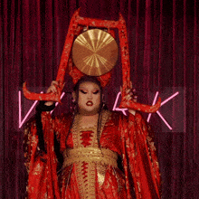 a woman in a red and gold dress is holding a large gong in front of a neon sign that says vilook